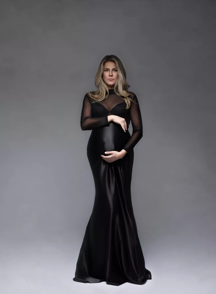 Glamorous maternity portrait with flowy dress and elegant posing - The Portrait Mama Elegant maternity portrait of a mother-to-be draped in fashion dress, standing against a soft-toned studio backdrop—capturing the grace and strength of motherhood.
